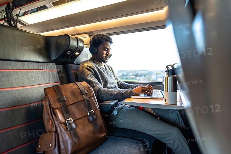 Young man wearing headphones working on a laptop while traveling by train, focusing on remote work and digital nomad lifestyle, enjoying a comfortable business trip