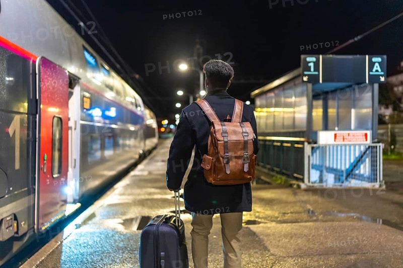 Man in coat and backpack pulls rolling luggage on a dim train platform at night, boarding for a business trip lone urban traveler heading into the city for work and adventure