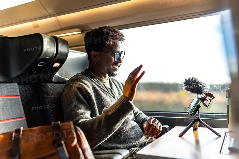 Man smiling and waving during a video call on a smartphone with a microphone on a tripod, traveling by train for business while working as a digital nomad influencer