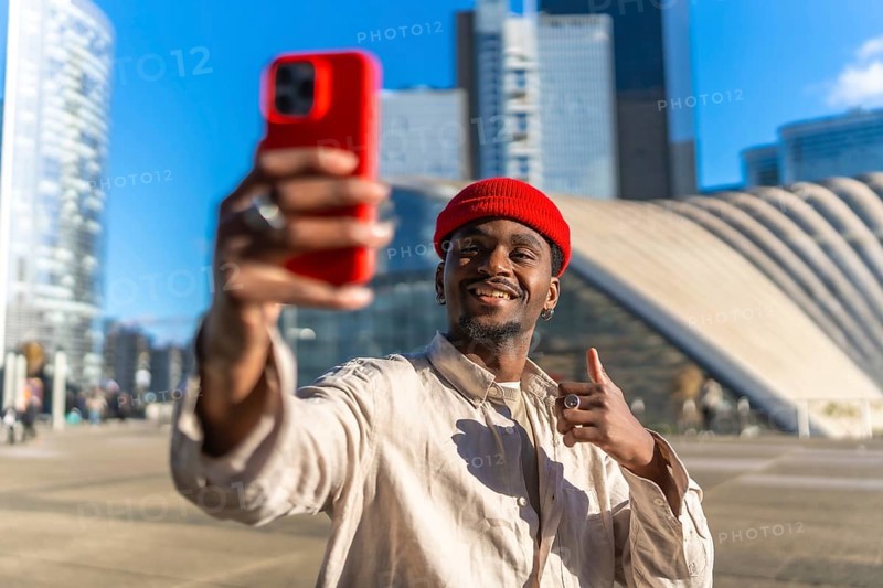 Young black man smiling while taking a selfie with a red smartphone and giving a thumbs up gesture, standing in front of modern architecture and skyscrapers under a blue sky