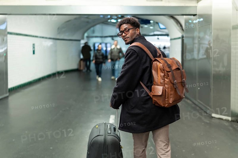Young man with a stylish leather backpack and rolling luggage is pausing to look back in a modern subway station or underground passage, preparing for his journey