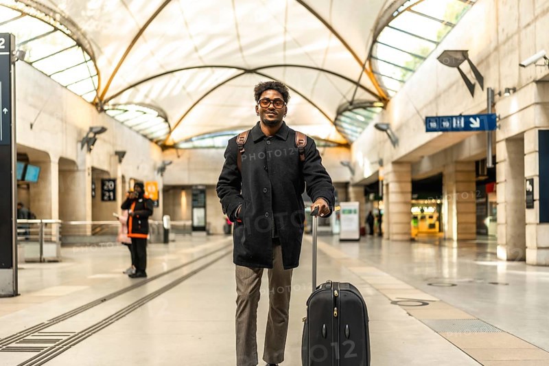 Young businessman standing on a modern train platform, smiling confidently with suitcase and backpack, ready for a city business trip and about to board his train