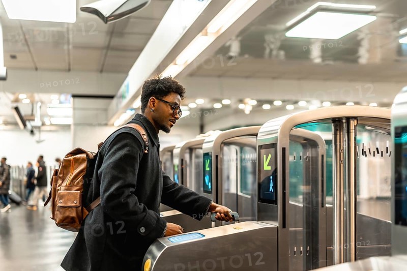 Young man with glasses and backpack tapping a contactless ticket at a modern subway turnstile, entering a bright underground station during his daily city commute