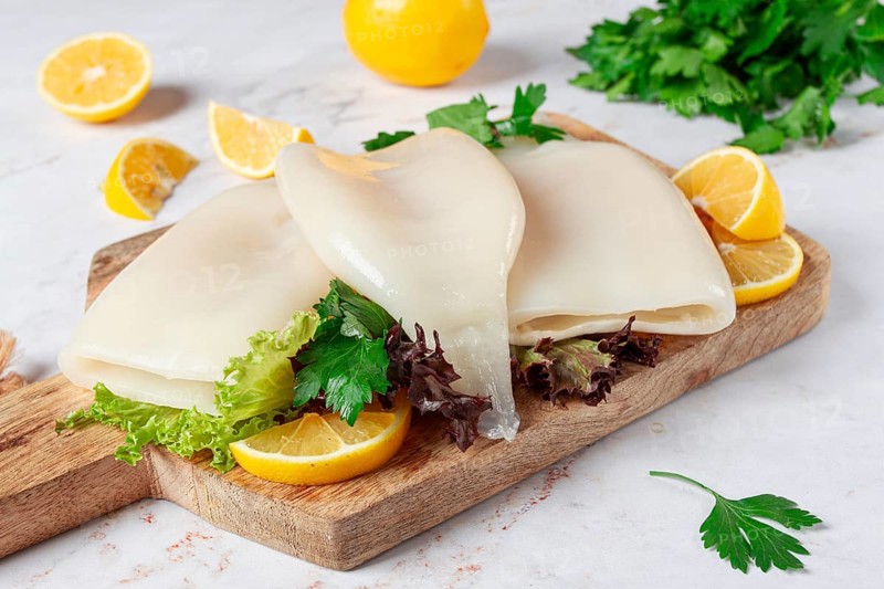 Fresh squid prepared for cooking, surrounded by herbs and lemon, placed on a wooden chopping board in a bright kitchen setting