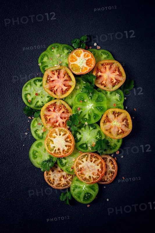 Sliced green tomatoes, on a dark surface, garnished with herbs and salt, top view