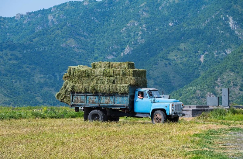 A blue truck with hay bales drives on a dirt road surrounded by green mountains, classic car, GAZ-53, Lorikeet province, Armenia