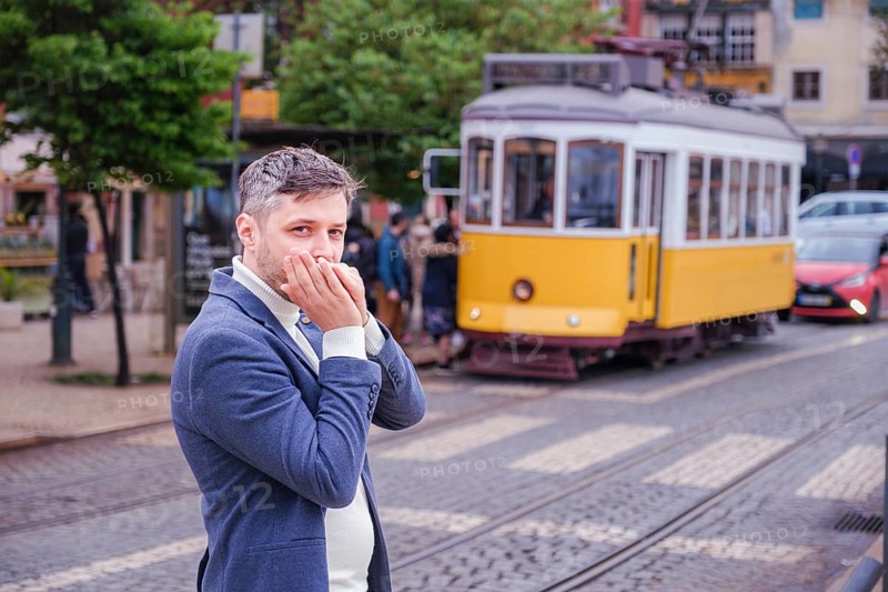 Man musician in a blue blazer and white sweater playing blues on a harmonica with eyes closed, standing outdoors in Lisbon city street, Portugal with tram in background