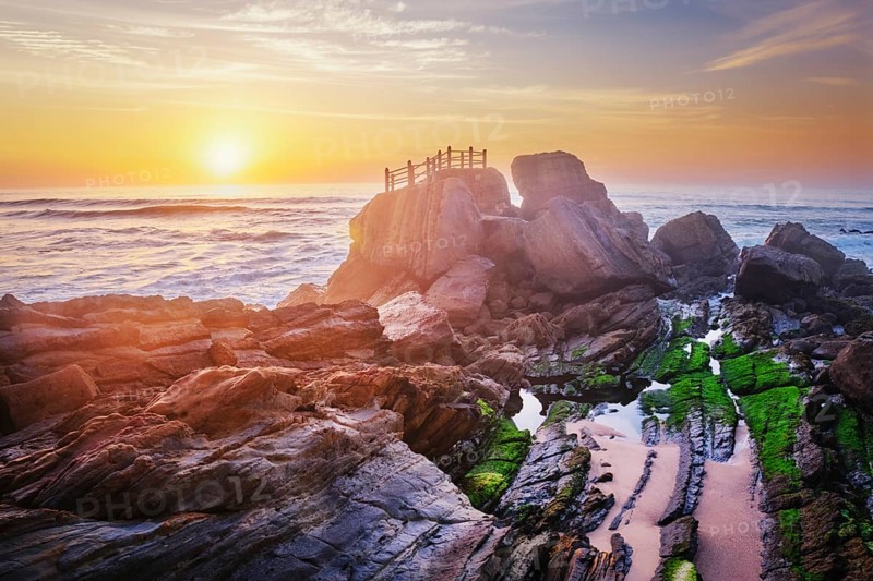 Scenic rock formation at Praia da Santa Cruz, Portugal, with ocean waves and vibrant sunset sky reflecting on the rocks