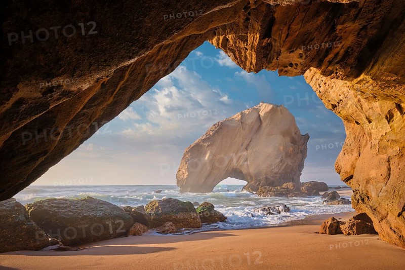Penedo do Guincho, a large boulder rock arch at Praia da Santa Cruz, Portugal, with ocean waves and sandy beach as seen from grotto cave on sunset