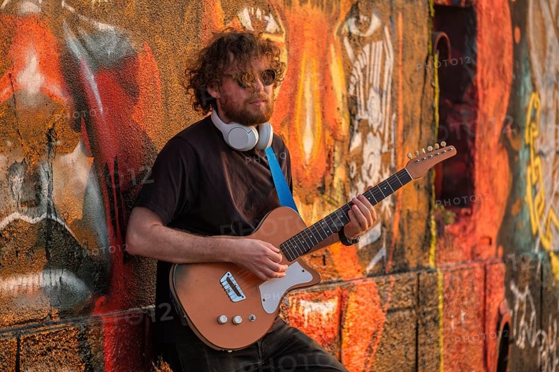 Hipster street musician in black playing electric guitar in the street on sunset leaning on a painted wall