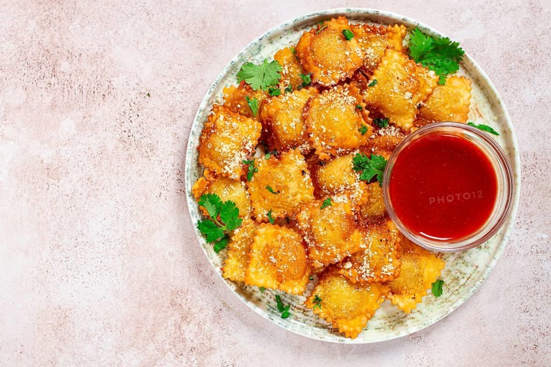 Fried ravioli with parmesan cheese, served with red sauce and garnished with herbs, on a light background, no people
