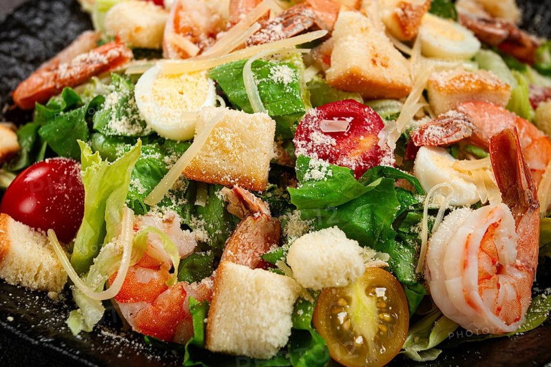 Caesar salad with tiger prawns, shrimp salad, lettuce leaves, quail eggs, cherry tomatoes, parmesan cheese, on a black plate