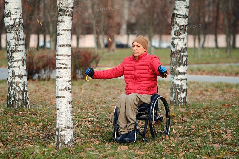 A senior man in wheelchair wearing red down jacket and gloves stretches arms outdoors in a grassy park among birch trees