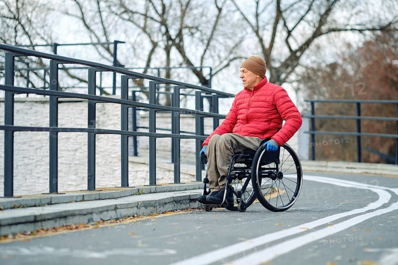 A senior man wearing a red jacket and hat is moving in his wheelchair along a smooth, winding path in a park. The trees are bare, indicating autumn, and he uses blue gloves for grip