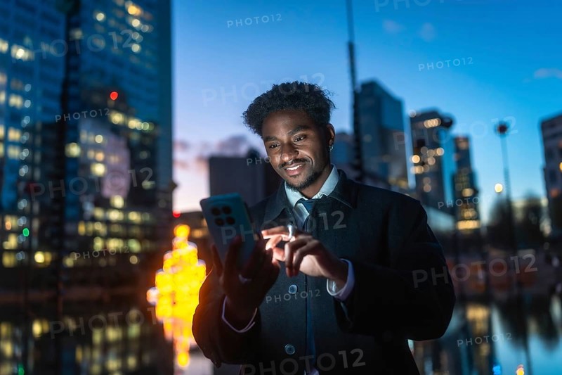 Young man standing outdoors at night, smiling while interacting with a smartphone, reflecting modern communication, global connectivity, and the digital lifestyle within a busy city environment