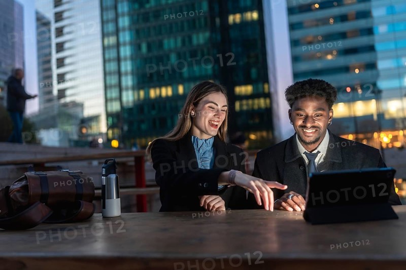 Two diverse business colleagues reviewing content on a digital tablet, discussing and laughing together after work hours in an urban setting with modern buildings