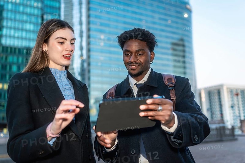 Two diverse business professionals collaborate outdoors in a modern city, smiling as they review data and strategy on a sleek tablet, sharing ideas and planning next steps