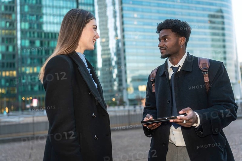 Two diverse business colleagues are engaging in a conversation about work, using a digital tablet while standing on a concrete bridge with modern glass skyscrapers in the urban background
