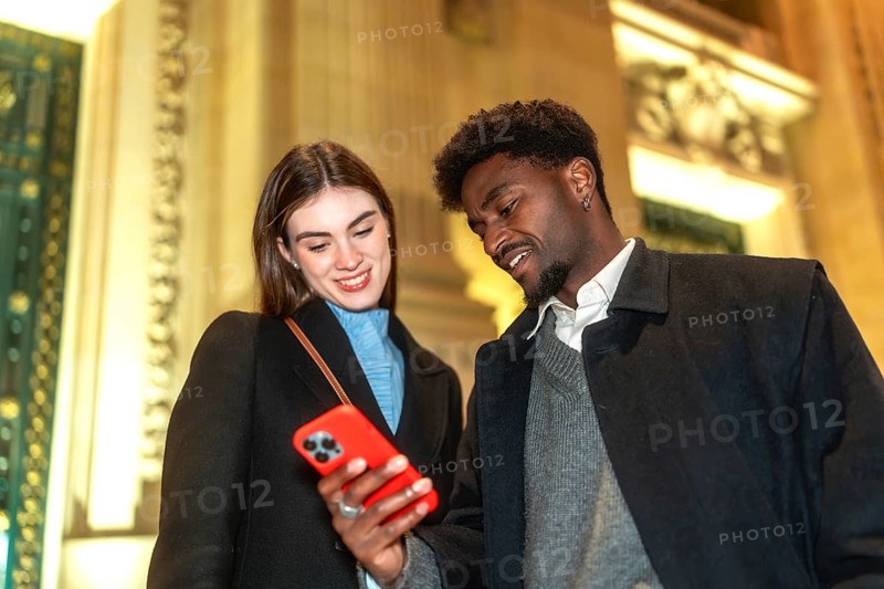 Diverse couple standing close, looking at a smartphone screen, smiling and connecting while sharing content on a modern device in an illuminated city environment at night