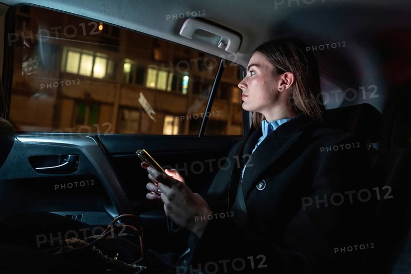 Woman traveling as a passenger in a ride hailing car at night, looking out the window while holding her illuminated smartphone, contemplating the urban cityscape passing by