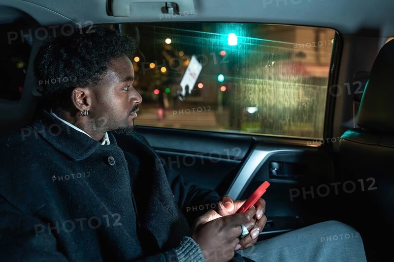 Afro american passenger riding in a car, looking out the window at city lights while holding a smartphone, capturing moments of urban public or private transport journey