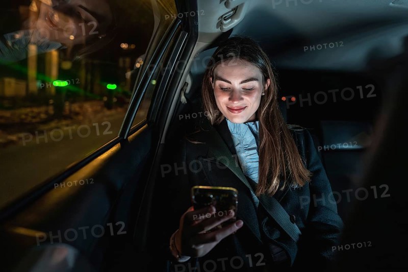 Young woman smiling while holding and looking at her smartphone in the backseat of a car at night, enjoying connected lifestyle during an urban journey
