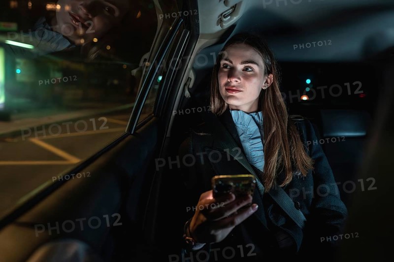 Woman travelling in the back seat of a car at night, holding a smartphone and looking out the window with city lights reflecting on the glass, representing urban transportation services