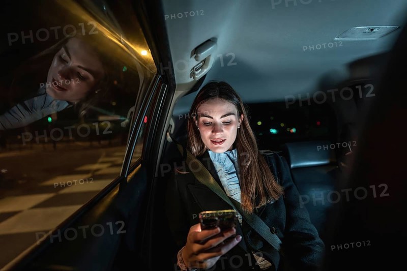 Young businesswoman in backseat at night, focused on smartphone screen while commuting through illuminated city streets, reflecting street lights and digital connection