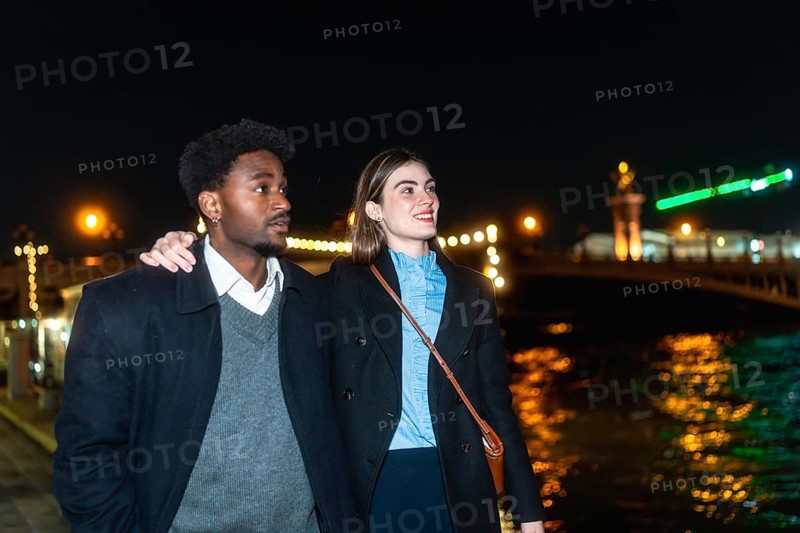 Young diverse couple strolling along a river promenade at night, city lights reflecting on the water as they hold each other, smiling and enjoying a romantic urban moment