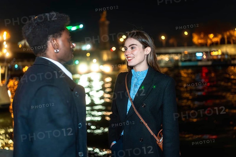 Young diverse couple standing by paris river at night, enjoying an intimate conversation during a romantic date, with city lights reflecting in the water creating a bokeh background
