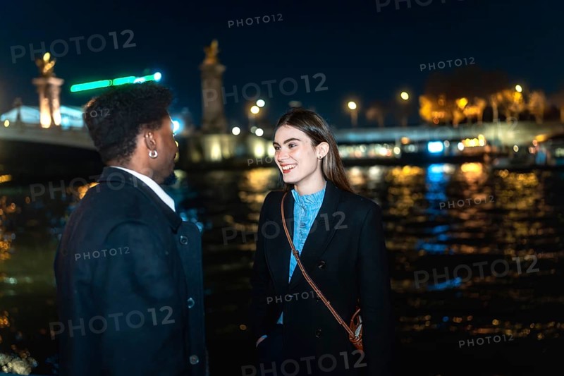 Young couple enjoying a romantic conversation by the illuminated river at night, sharing an intimate moment in the vibrant city lights of paris, france