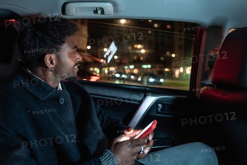 African american man in backseat of car at night smiling as he holds a glowing smartphone, looking out the window amid city lights and blurred motion during a relaxed ride share journey