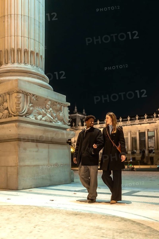 Interracial couple strolling arm in arm past an illuminated historic monument at night, sharing a romantic, candid moment of conversation and connection in the city