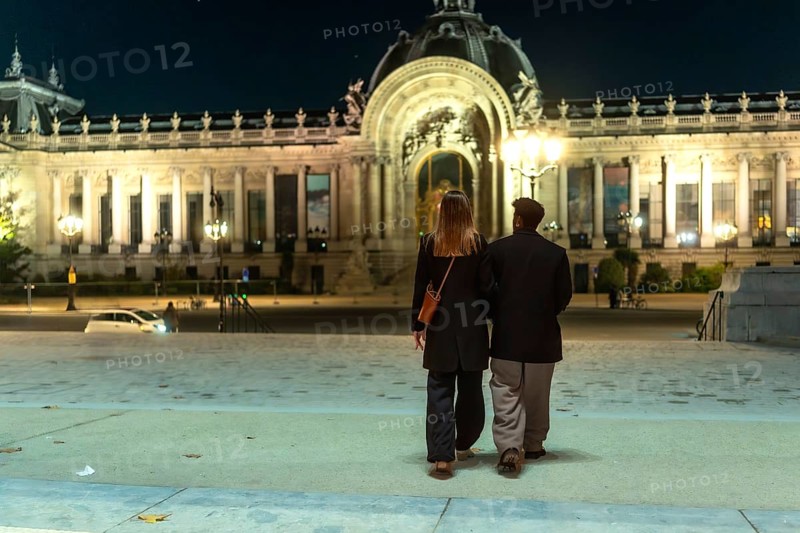 Couple walking arm in arm beneath the illuminated facades of petit palais at night, enjoying a romantic evening stroll along grand parisian streets, architecture glowing in warm light