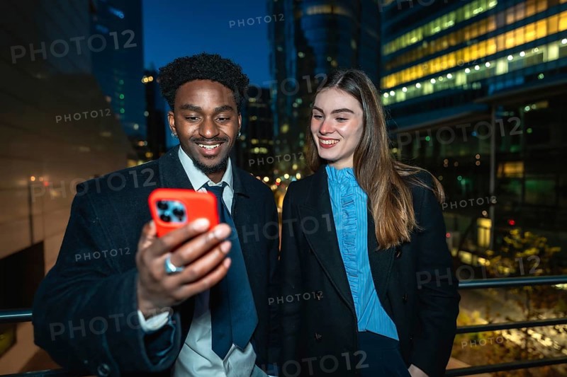 Diverse business colleagues smiling and looking at a smartphone together, enjoying a moment while connecting and sharing digital content against a modern city backdrop at night