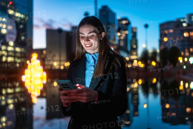 Young professional woman smiling happily while interacting with her mobile phone against a blurred city background with bokeh lights and reflections in the evening