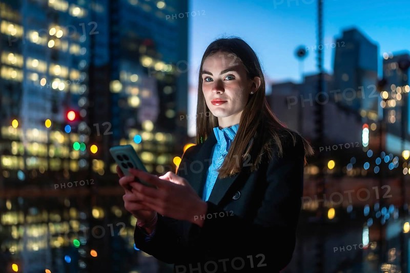 Young businesswoman looking at camera while holding a mobile phone in her hands and texting, standing in an urban environment with illuminated buildings at dusk
