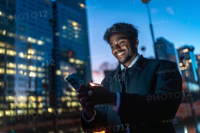 Young black man connecting with his smartphone, illuminated by the screen's glow, against a blurred backdrop of modern city buildings at dusk, representing urban technology and communication