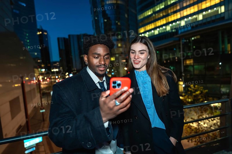 Diverse business professionals standing outside in a city at night, sharing content on a smartphone, connecting through technology and networking in an urban setting