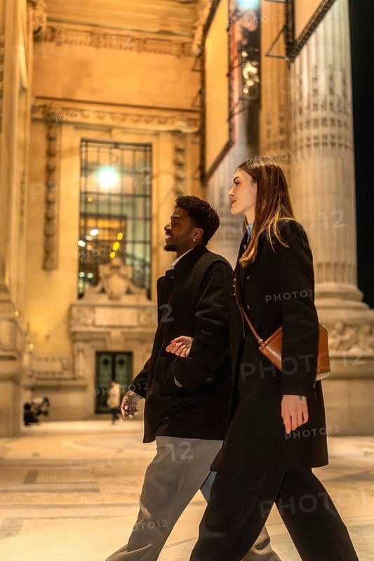 Diverse couple walking arm in arm on an illuminated city street at night, exploring urban architecture and enjoying a romantic evening stroll in downtown