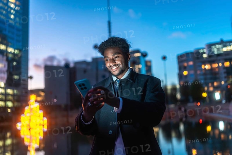 African american man smiling while engaging with a smartphone, viewing content under the illuminated city lights at night, embodying urban connectivity and modern communication