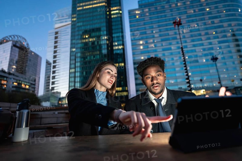 Diverse business colleagues working together on a digital tablet, pointing at the screen, with modern urban skyscrapers illuminating the evening sky in the background