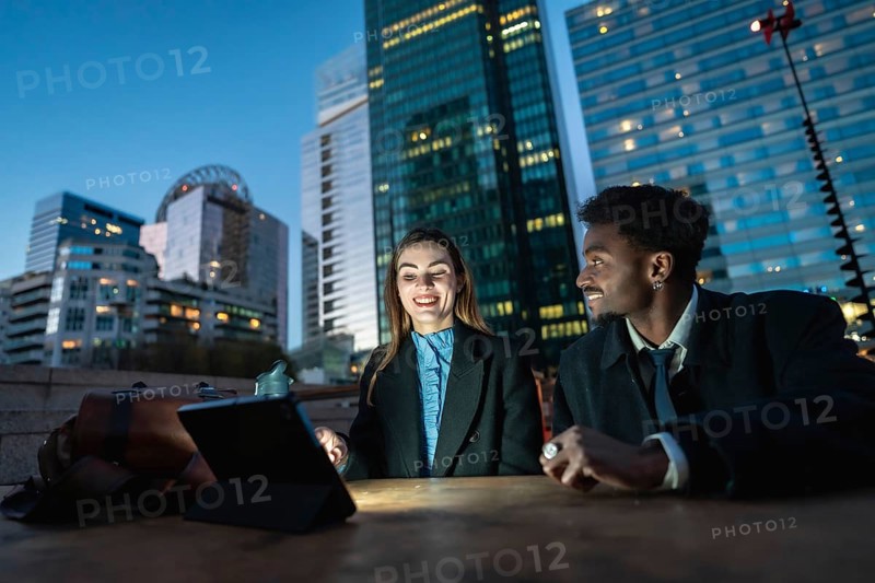 Two diverse business colleagues share a tablet at an outdoor table with a glowing downtown skyline at dusk, collaborating on digital strategy, finance and global communication