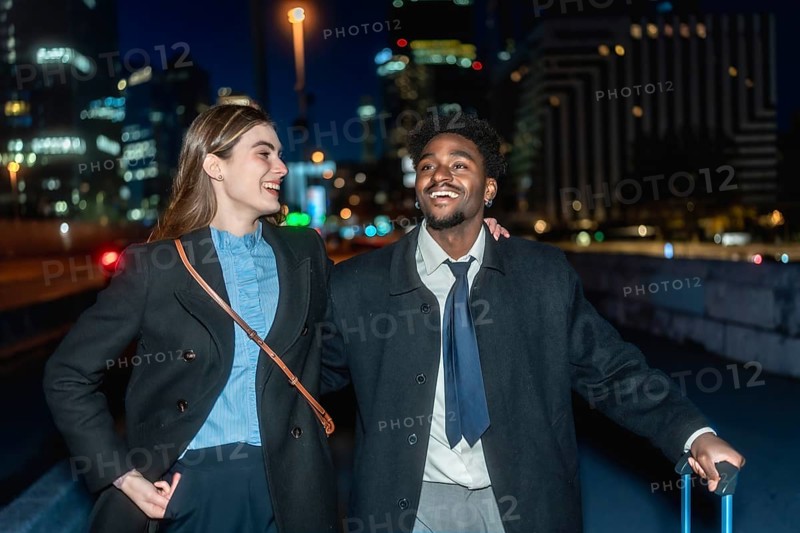 Diverse business colleagues smiling and walking together carrying luggage in an urban setting at night, representing successful travel, partnership, and global business connections