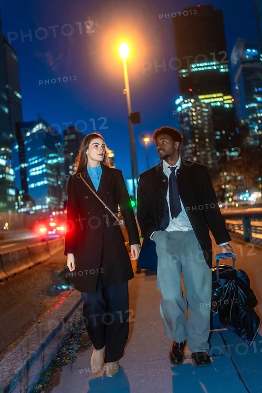 Diverse business couple walking with luggage along a downtown sidewalk at night, passing illuminated skyscrapers and a street lamp as they arrive or depart on a city trip