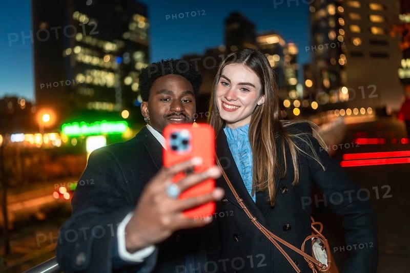 Diverse friends posing for a self portrait with a smartphone, smiling warmly as they enjoy a casual evening outing in the vibrant, illuminated urban cityscape