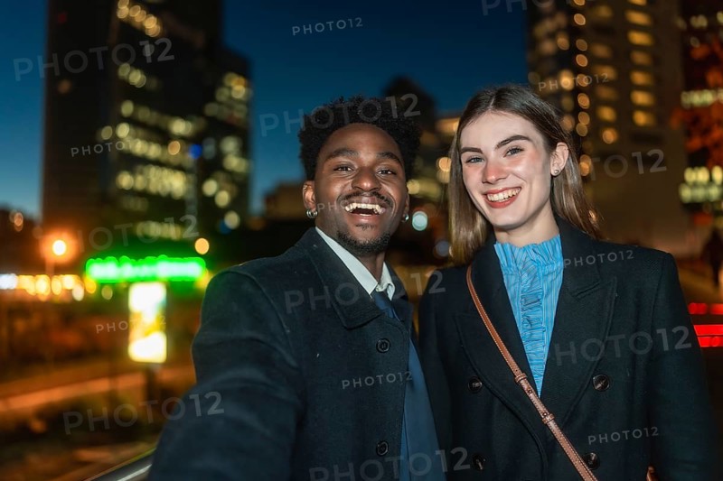 Diverse young man and woman smiling happily while taking a selfie together outdoors at night, with a modern glowing city skyline creating a soft bokeh background