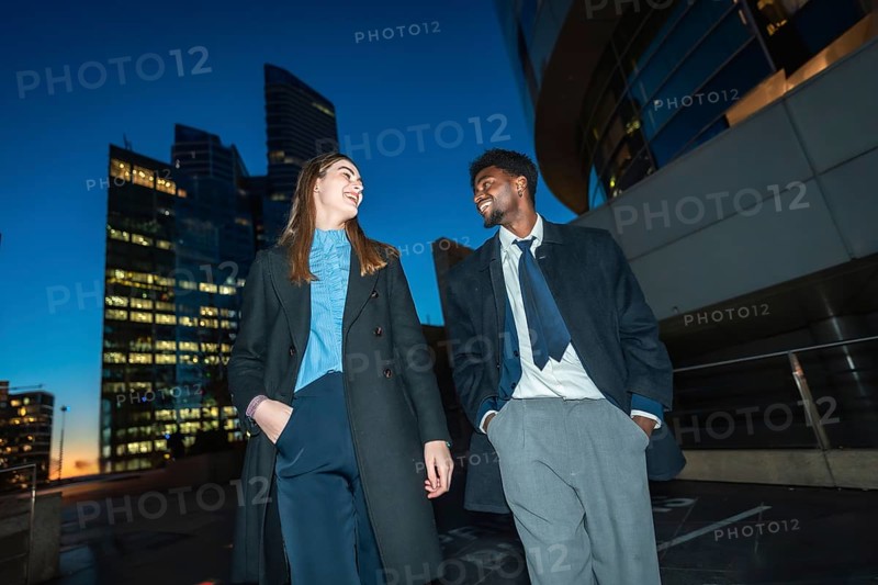Diverse business colleagues walking down a city street at night, smiling and laughing together with modern office buildings providing a vibrant urban backdrop