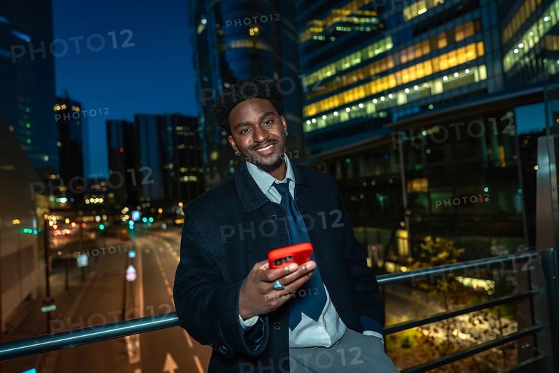 Young black businessman standing on a bridge, holding a modern smartphone with a red case, smiling towards the camera against a backdrop of illuminated city buildings and traffic lights at night