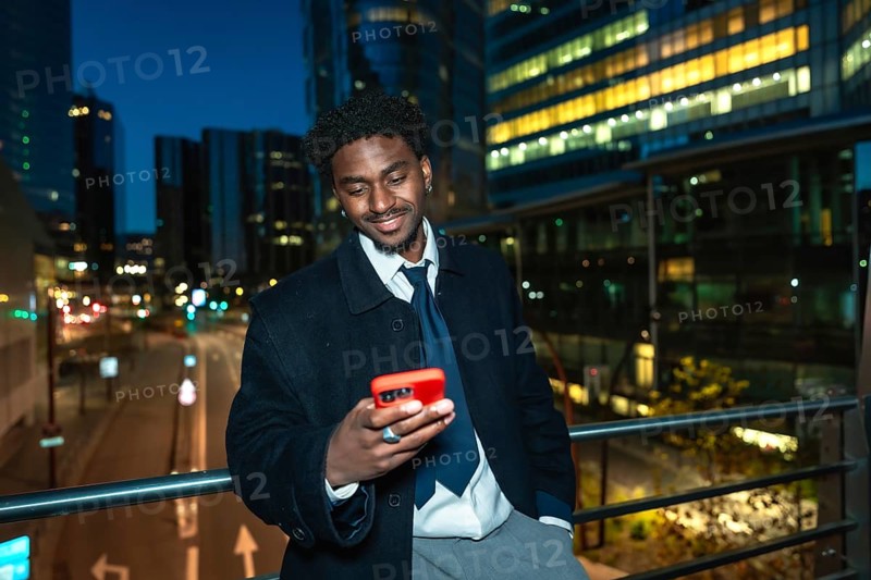 Young black businessman on an elevated walkway at night, smiling as he uses his smartphone with blurred city lights and modern buildings behind, conveying urban tech and connection